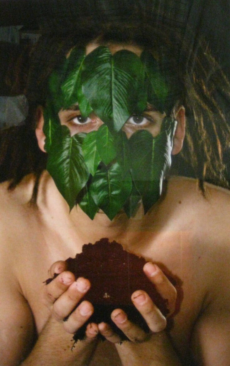 Color photograph of a person with their face covered in bright green leaves, no shirt, hands full of dirt.